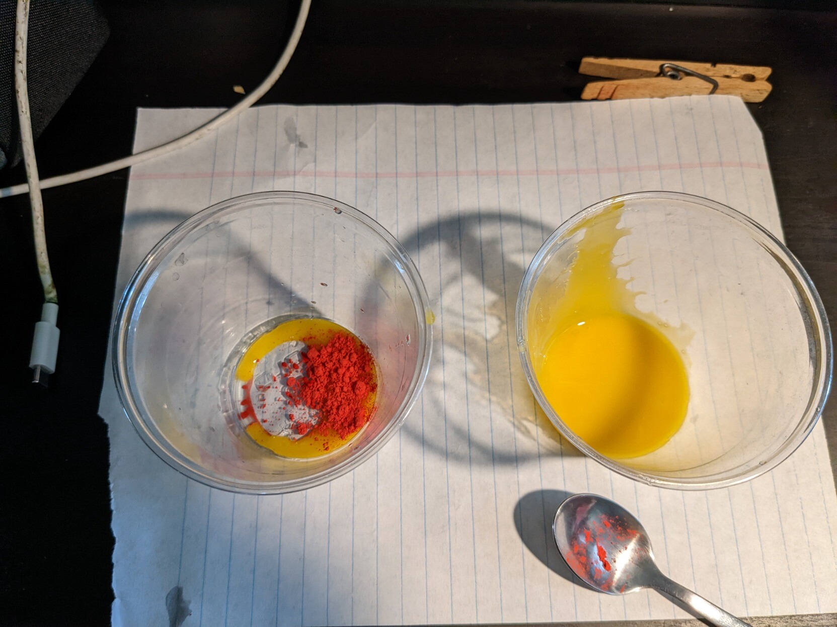 Two plastic cups with a broken egg yolk in each. In one of them is a red powder, which is bleeding in a bit in a kind of sunset-y way.